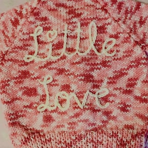 Könnte beinhalten: Ein gestrickter Pullover in Rosa- und Rottönen mit den Worten "Little Love" aus weißem Garn gestickt. Der Pullover hat ein strukturiertes Muster und ein Herz über dem Wort "Little".