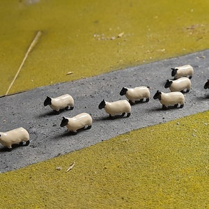 以下が含まれることがあります： 灰色の道路を歩くおもちゃの羊の群れを描いたミニチュアシーン。羊はクリーム色の体と黒い頭と脚をしています。1匹の羊は茶色です。道路は緑と黄色の地形に囲まれています。