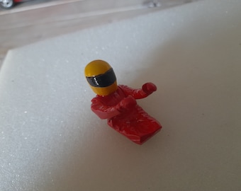 May include: A red and yellow toy race car driver figurine with a helmet. The driver is in a racing position with arms outstretched.