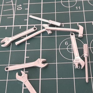 Könnte beinhalten: Eine Sammlung von silbernen Miniaturwerkzeugen, darunter Schraubenschlüssel, ein Hammer und Schraubendreher, ist auf einer grünen Schneidematte angeordnet. Die Werkzeuge sind detailliert und scheinen aus Metall zu sein.