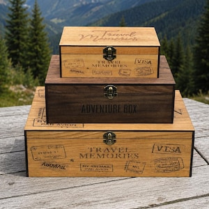 May include: Three rectangular wooden boxes stacked on top of each other. The top and bottom boxes are light wood with black travel stamp graphics and the text Travel Memories. The middle box is dark wood with the text Adventure Box engraved on the front.