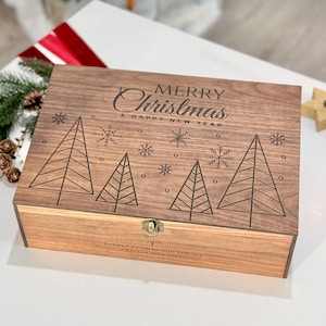 Könnte beinhalten: Weihnachtsgeschenkbox aus Holz mit Scharnierdeckel. Der Deckel ist mit den Worten "MERRY Christmas & HAPPY NEW YEAR", Weihnachtsbäumen und Schneeflocken graviert. Die Box wird mit einem Metallverschluss verschlossen.
