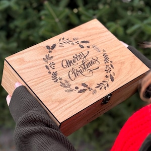 Peut inclure: Une boîte en bois avec un message "Merry Christmas" gravé au laser, entouré d'une couronne. La boîte est tenue par une personne portant un pull vert foncé et une écharpe rouge. L'arrière-plan est un arbre vert flou.