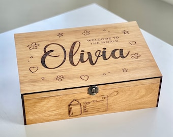 May include: A wooden keepsake box with a hinged lid. The lid is engraved with the text "Welcome to the world Olivia" and features a floral design. The box has a latch and a small engraved image of a baby bottle on the front.