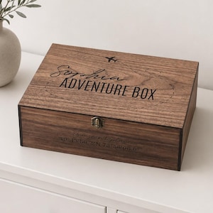 Peut inclure: Une boîte rectangulaire en bois foncé avec un couvercle à charnière et un loquet en métal. Le couvercle présente le texte gravé "Sophia ADVENTURE BOX" avec une icône d'avion et une trajectoire de vol en pointillés. La face avant porte le texte "Our secret place" et des coordonnées.