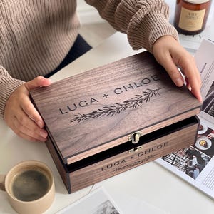 May include: A wooden box with the engraved text "LUCA + CHLOE" and a decorative floral design. The box is open, revealing a black interior. A small metal clasp secures the lid. A cup of coffee and a candle are also visible.