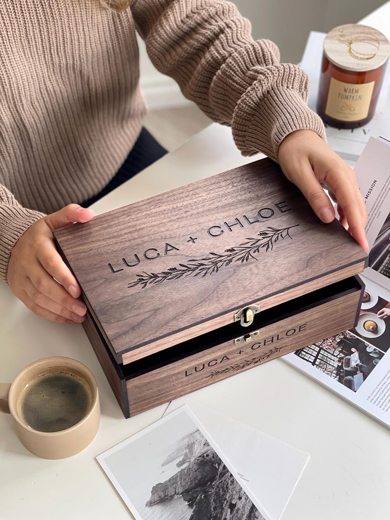 May include: A wooden box with the engraved text "LUCA + CHLOE" and a decorative floral design. The box is open, revealing a black interior. A small metal clasp secures the lid. A cup of coffee and a candle are also visible.