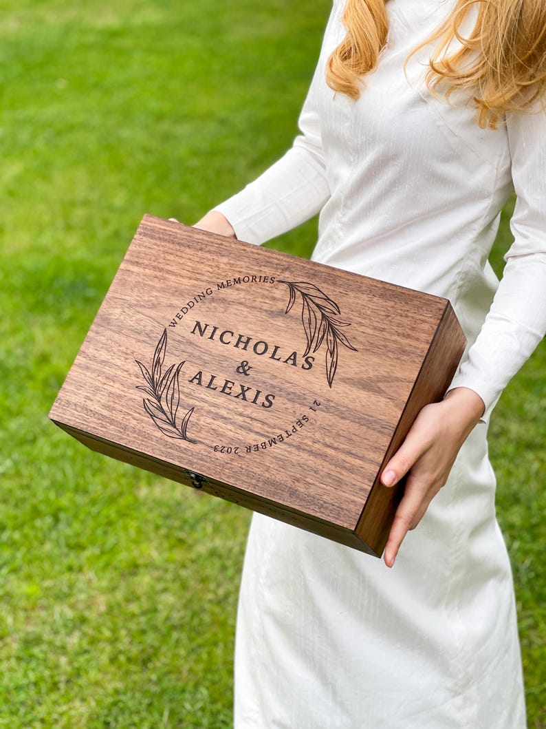 Pu&ograve; includere: Un cofanetto in legno marrone scuro con la scritta incisa "NICHOLAS & ALEXIS" all'interno di una ghirlanda floreale e la data "21 SETTEMBRE 2023". Il cofanetto &egrave; etichettato "WEDDING MEMORIES".