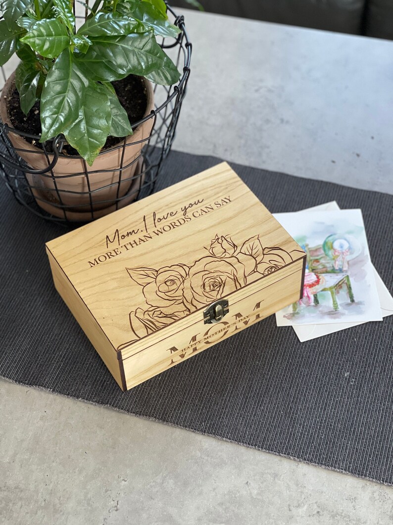 Puede incluir: Una caja de madera con tapa con bisagras y cierre de lat&oacute;n. La caja est&aacute; grabada con el texto "Mam&aacute;, te amo m&aacute;s de lo que las palabras pueden decir" y un dise&ntilde;o floral. La caja tambi&eacute;n tiene el texto "Feliz D&iacute;a de la Madre Mam&aacute;" grabado en la parte inferior.