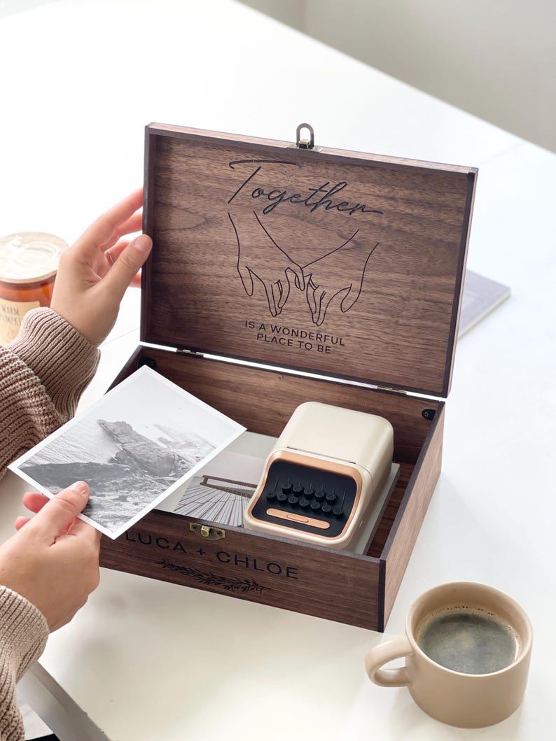 May include: A wooden keepsake box with the word "Together" and a line drawing of two hands holding each other. The box contains a small beige and gold label maker, a black and white photo, and is labeled "LUCA + CHLOE". A cup of coffee sits nearby.