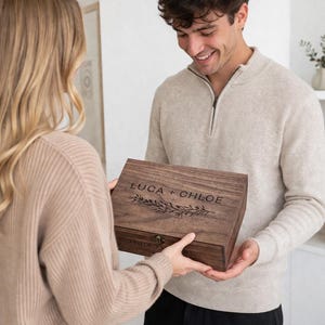 May include: A wooden box with the names "LUCA + CHLOE" engraved on the lid, along with a decorative leaf design. The box is being held by two people. The box has a clasp closure.