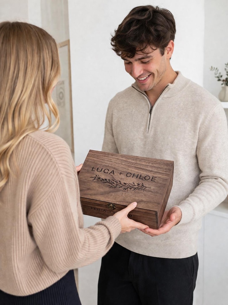 May include: A wooden box with the names "LUCA + CHLOE" engraved on the lid, along with a decorative leaf design. The box is being held by two people. The box has a clasp closure.