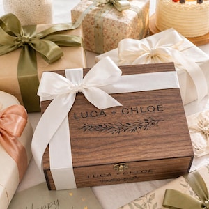 May include: A wooden box with the text "LUCA + CHLOE" and a floral design, surrounded by wrapped gifts with ribbons and bows. A birthday cake with berries and a lit candle is also present, along with a card that says "Happy Birthday!"