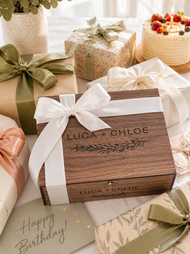 May include: A wooden box with the text "LUCA + CHLOE" and a floral design, surrounded by wrapped gifts with ribbons and bows. A birthday cake with berries and a lit candle is also present, along with a card that says "Happy Birthday!"