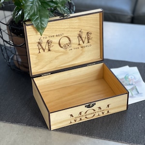 Puede incluir: Caja de madera con tapa con bisagras. La tapa est&aacute; abierta y el interior de la caja es visible. La caja est&aacute; decorada con las palabras "To the Best Mom in the World" y "Happy Mother's Day Mom".