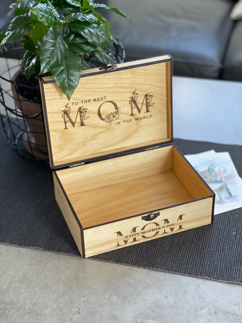 Puede incluir: Caja de madera con tapa con bisagras. La tapa est&aacute; abierta y el interior de la caja es visible. La caja est&aacute; decorada con las palabras "To the Best Mom in the World" y "Happy Mother's Day Mom".