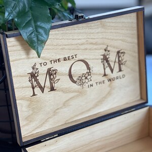 Puede incluir: Una caja de madera con tapa con bisagras. La tapa est&aacute; abierta y el interior de la caja es visible. La tapa tiene un mensaje grabado que dice "To the best Mom in the world" con acentos florales alrededor de las letras.