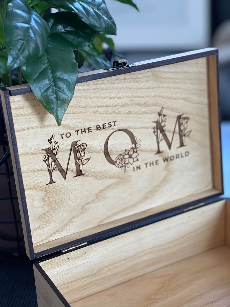 Puede incluir: Una caja de madera con tapa con bisagras. La tapa est&aacute; abierta y el interior de la caja es visible. La tapa tiene un mensaje grabado que dice "To the best Mom in the world" con acentos florales alrededor de las letras.