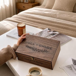 May include: A dark wooden box with the names "LUCA + CHLOE" engraved on the lid, along with a floral design. A small, beige coffee cup sits on a white table. The box is closed with a small clasp.