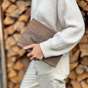 May include: A rectangular wooden plaque with the name "LUCA" engraved in a serif font, accompanied by a sprig design. The plaque is a dark brown color and appears to be held by a person. The background is a stack of firewood.