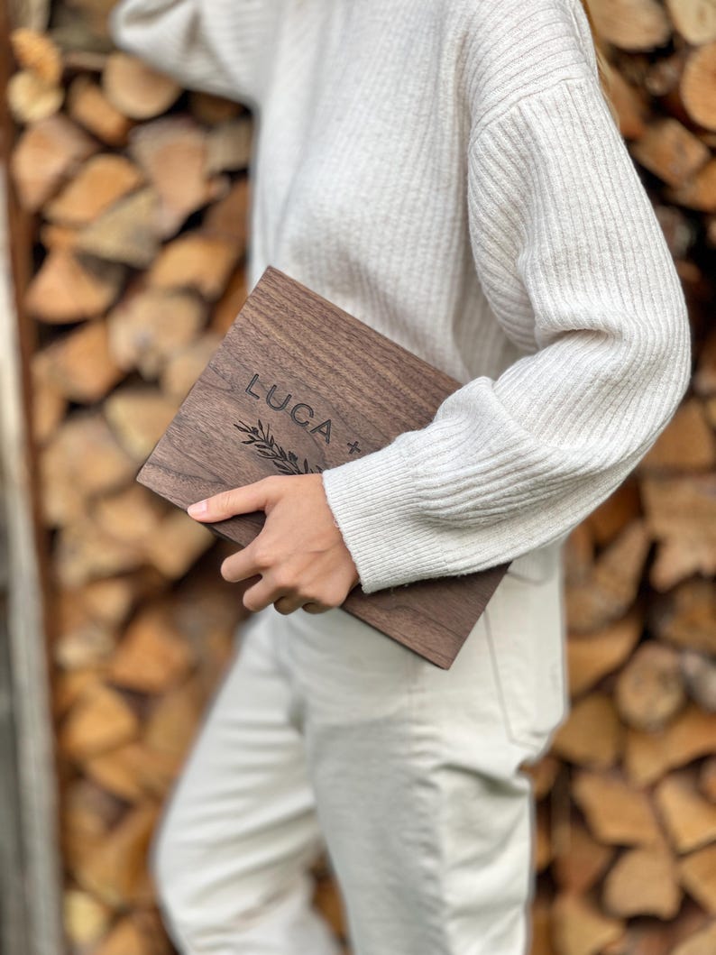 May include: A rectangular wooden plaque with the name "LUCA" engraved in a serif font, accompanied by a sprig design. The plaque is a dark brown color and appears to be held by a person. The background is a stack of firewood.