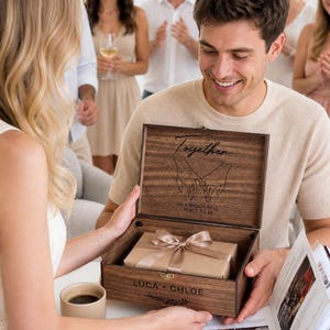 May include: A wooden keepsake box with a gift inside, tied with a satin bow. The box is engraved with the word "Together" and the names "Luca + Chloe". The box is being opened by a person, with a group of people in the background.
