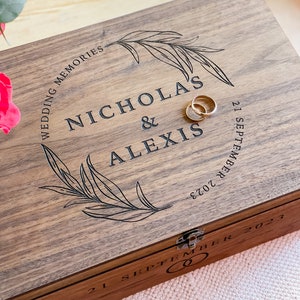Pu&ograve; includere: Un cofanetto in legno con i nomi "NICHOLAS & ALEXIS" incisi sul coperchio, insieme alla data "21 SETTEMBRE 2023". Il cofanetto &egrave; decorato con un motivo a foglie circolare e due fedi nuziali d'oro.