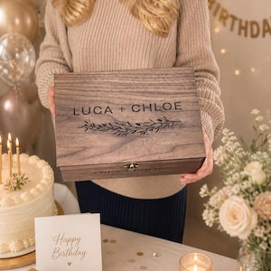 May include: A wooden box with the names "LUCA + CHLOE" engraved on the lid, along with a floral design. The box is held in front of a birthday scene with a cake, a "Happy Birthday" sign, and a gift box.