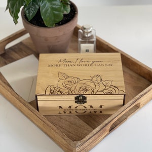 Puede incluir: Caja de madera con tapa con bisagras y cierre de lat&oacute;n. La caja est&aacute; grabada con las palabras "Mam&aacute;. Te amo m&aacute;s de lo que las palabras pueden decir." y un dise&ntilde;o floral. La caja tambi&eacute;n tiene las palabras "Feliz D&iacute;a de la Madre Mam&aacute;" grabadas en la parte inferior.