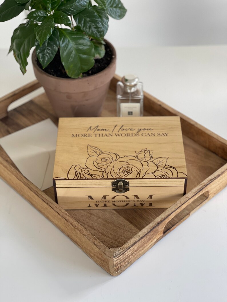 Puede incluir: Caja de madera con tapa con bisagras y cierre de lat&oacute;n. La caja est&aacute; grabada con las palabras "Mam&aacute;. Te amo m&aacute;s de lo que las palabras pueden decir." y un dise&ntilde;o floral. La caja tambi&eacute;n tiene las palabras "Feliz D&iacute;a de la Madre Mam&aacute;" grabadas en la parte inferior.
