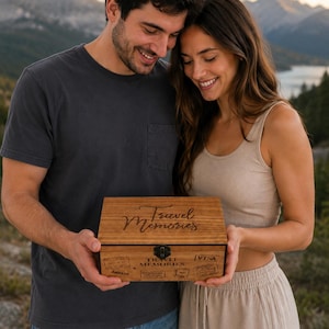 Puede incluir: Una caja rectangular de madera con tapa abatible y cierre metálico. La tapa presenta el texto grabado Travel Memories en letra cursiva. El panel frontal muestra el texto Travel Memories y varios sellos temáticos de viajes.