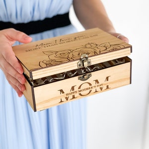 Puede incluir: Una caja de madera con tapa abatible. La tapa est&aacute; abierta y muestra un dise&ntilde;o floral con el texto "Mam&aacute;, te quiero m&aacute;s de lo que las palabras pueden decir". La parte inferior de la caja tiene el texto "Mam&aacute; Feliz D&iacute;a de la Madre".