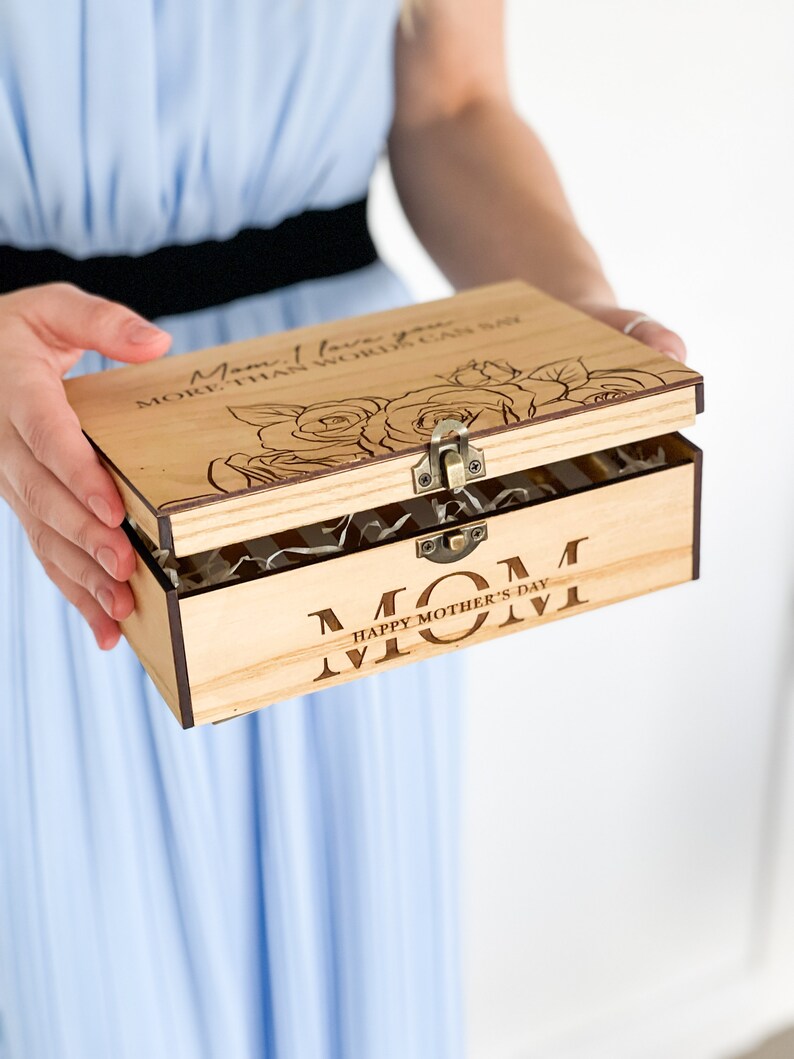 Puede incluir: Una caja de madera con tapa abatible. La tapa est&aacute; abierta y muestra un dise&ntilde;o floral con el texto "Mam&aacute;, te quiero m&aacute;s de lo que las palabras pueden decir". La parte inferior de la caja tiene el texto "Mam&aacute; Feliz D&iacute;a de la Madre".
