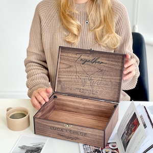 May include: A wooden keepsake box with the lid open, revealing the interior. The lid is engraved with the word "Together" and a graphic of two hands holding each other. The box also has the names "LUCA + CHLOE" engraved on the front.