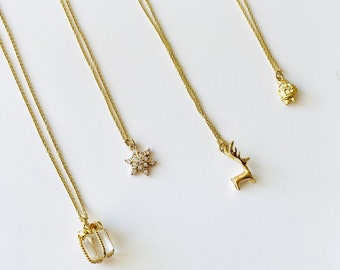 May include: Four gold necklaces with different charms. One necklace has a snowflake charm, one has a reindeer charm, one has a small box charm, and one has a small geometric charm.