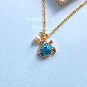 May include: A gold-colored necklace featuring a small turtle pendant with blue and turquoise enamel detailing. A small pearl is also attached to the chain. The necklace is displayed on a light blue surface.
