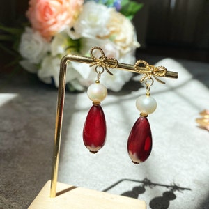 May include: A pair of dangle earrings with a red teardrop-shaped stone and a white pearl bead. The earrings are attached to a gold bow.