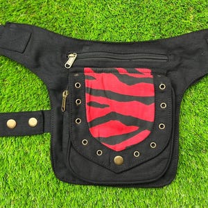 Puede incluir: Riñonera negra con un bolsillo con estampado de cebra rojo y negro. La riñonera tiene un compartimento principal con cremallera, un bolsillo más pequeño con cremallera y una correa con cuatro cierres de botón. La riñonera está sobre un fondo verde.