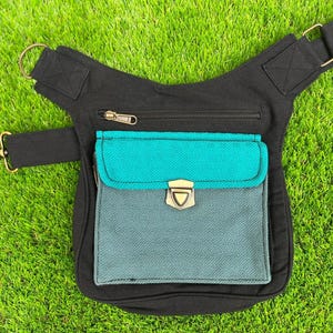 Puede incluir: Riñonera negra con un bolsillo delantero turquesa y gris. La bolsa tiene una cremallera y un cierre dorados. El bolsillo tiene un cierre dorado. La bolsa está sobre una superficie de hierba verde.