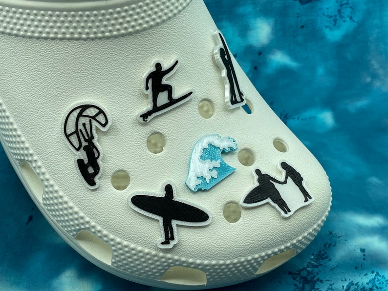 May include: White clogs with various black and blue themed shoe charms. The charms include a surfer, a wave, a kite surfer, and a couple holding hands on a surfboard.