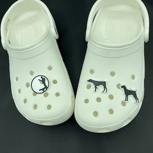 May include: A pair of white clogs with black dog and hunter silhouette charms. The charms are attached to the front of the clogs.