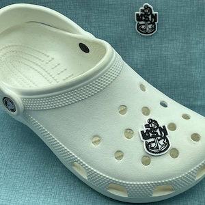 May include: White Crocs shoe with a black and white USN anchor charm attached. A second charm is visible nearby. The shoe has multiple ventilation holes and a textured surface. The background is a light blue.