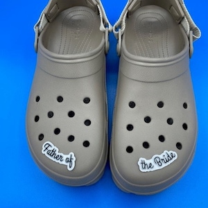 May include: A pair of tan-colored clogs with a strap on the back. The left clog has a white sticker that says "Father of" and the right clog has a white sticker that says "the Bride".
