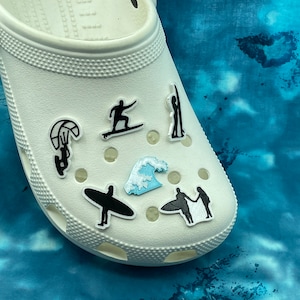May include: White clogs with various black and blue stickers depicting different water sports, including surfing, kiteboarding, and a wave.