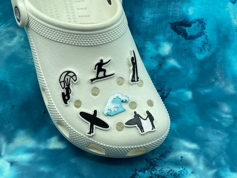 May include: White clogs with various black and blue stickers depicting different water sports, including surfing, kiteboarding, and a wave.