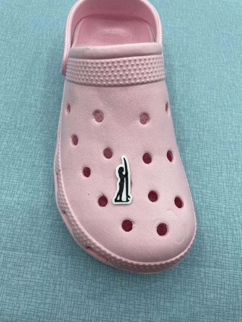 May include: Pink Crocs with a black and white silhouette of a person surfing on a surfboard.