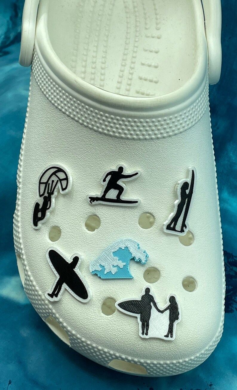 May include: White shoe with various black and blue silhouette charms of people surfing, kiteboarding, and riding waves.