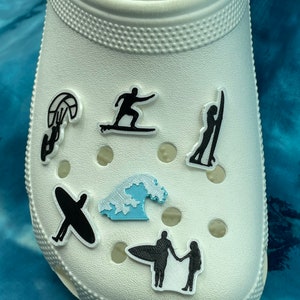 May include: White shoe with black and blue charms. The charms are of a surfer, a wave, a kite surfer, a person holding a surfboard, and a person surfing.