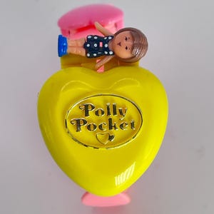 May include: A vintage Polly Pocket compact in the shape of a yellow heart with a gold-colored border. The compact has a small doll figure on top, wearing a blue and white polka dot outfit. The compact is a collectible toy.