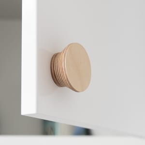 Pode incluir: Close-up de uma maçaneta de armário de madeira com um design em camadas. A maçaneta é de cor marrom claro e circular, e está montada em uma porta de armário branca.